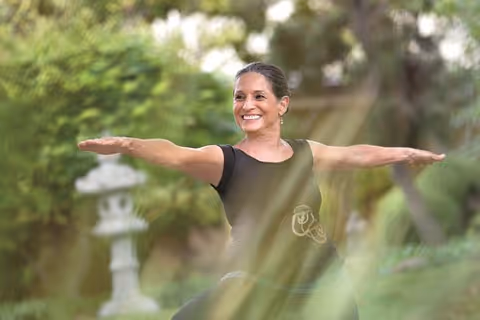A woman outdoors practicing yoga in a green garden setting, smiling with arms extended in a yoga pose.