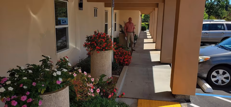 Outdoor covered walkway at Village Oaks Senior Care with large flower pots filled with colorful flowers along the side. A man in a red shirt and beige pants is standing near the end of the walkway. Several parked cars are visible on the right side.