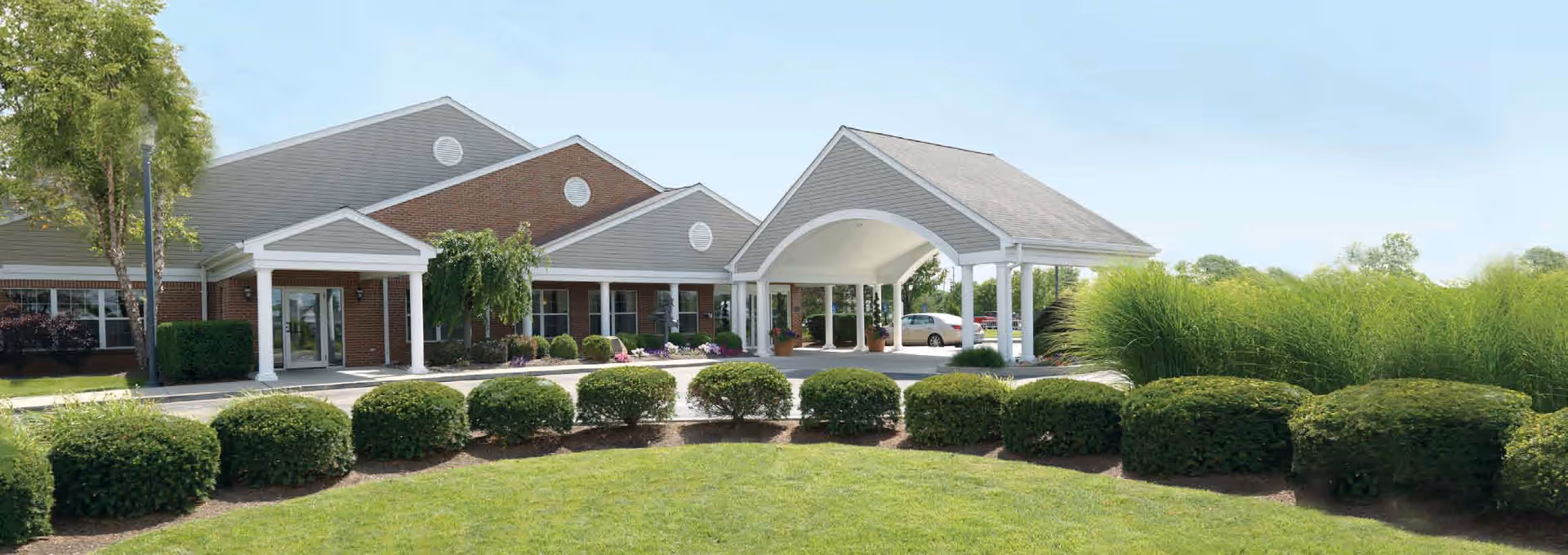 Exterior view of Dayspring Skilled Nursing Facility showing a brick building with a covered entrance supported by white columns, surrounded by neatly trimmed bushes and green grass under a clear sky.