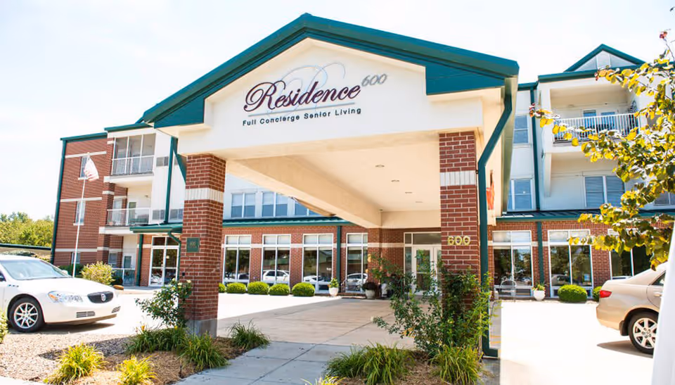 Entrance of Residence 600, a senior living facility with a covered drop-off area supported by brick columns, surrounded by a multi-story building with balconies and large windows. Cars are parked nearby and landscaping includes bushes and small trees.