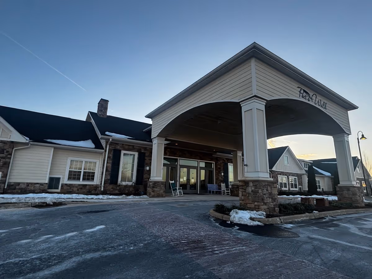Covered main entrance and porte-cochere of a senior living building with stone pillars, rocking chairs, and light snow on the ground.