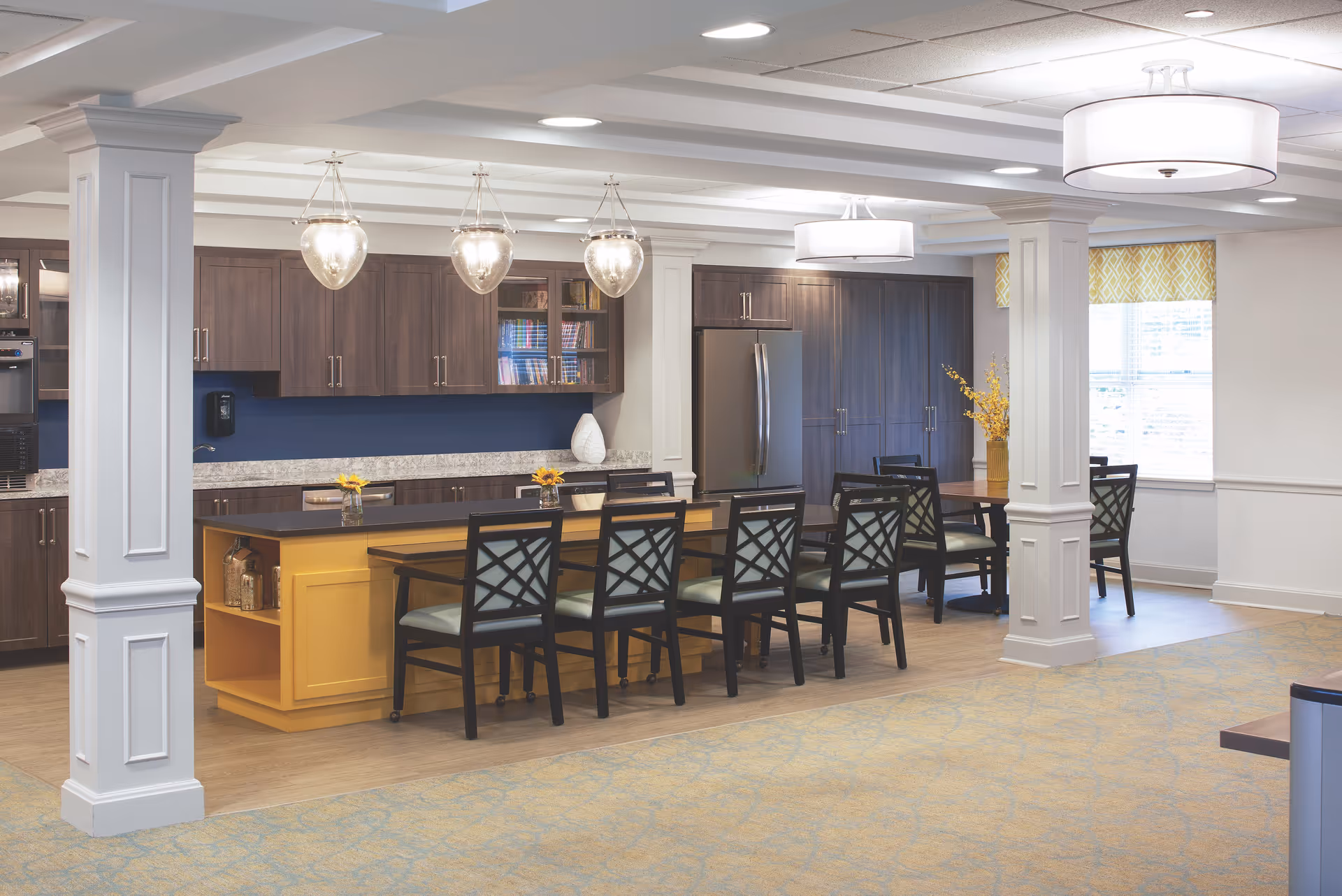 Open communal kitchen and dining area with a long yellow island, dark wood cabinets, pendant lights, and several chairs.
