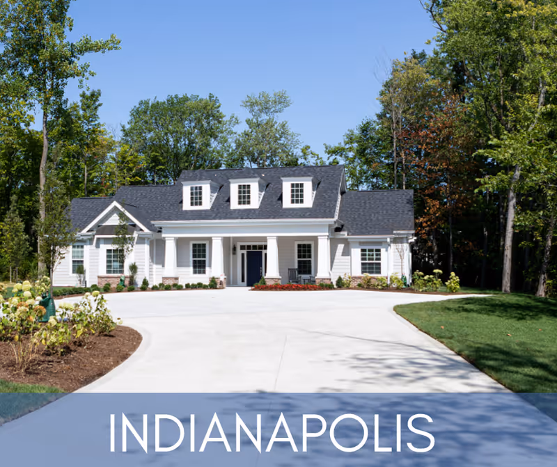 Front view of a white cottage-style building with columns, dormer windows, a wide driveway, and surrounding trees.