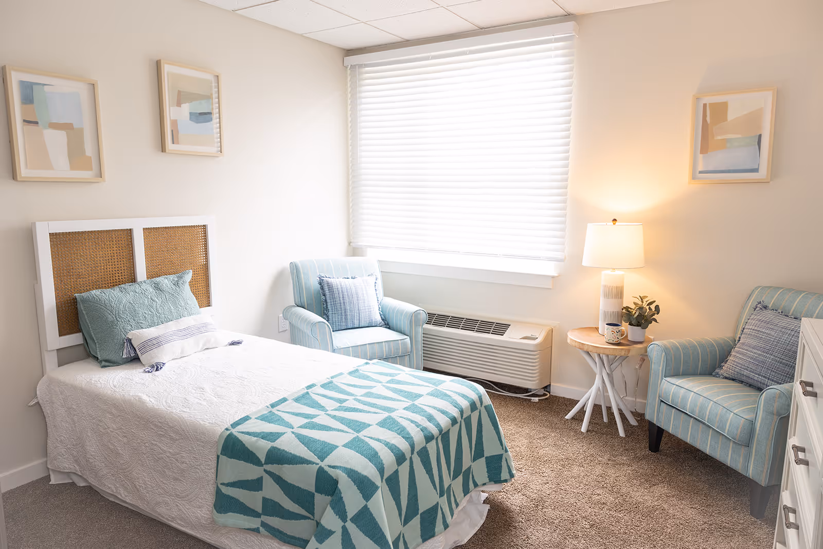 Bright bedroom with a single bed, teal patterned throw, two upholstered chairs, a side table with lamp, and a large window.