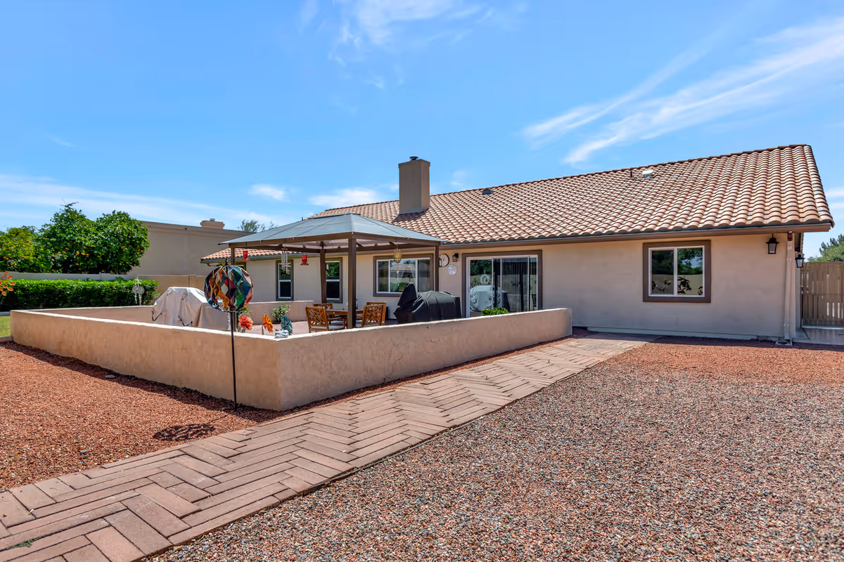 Outdoor patio area of a single-story building with a tiled roof under a clear blue sky. The patio is enclosed by a low beige wall and features a covered seating area with a table and chairs, a barbecue grill, and decorative garden ornaments. The ground is covered with reddish gravel and a paved walkway leads to the patio.