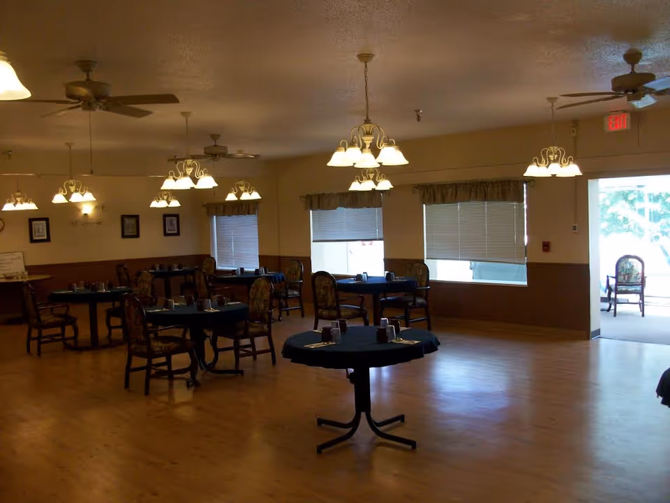 Interior view of a dining room with several round tables covered with dark tablecloths and set with napkins and condiments. The room has wooden floors, multiple ceiling fans, and hanging light fixtures. Windows with blinds and valances line one wall, and an exit door is visible on the right side.