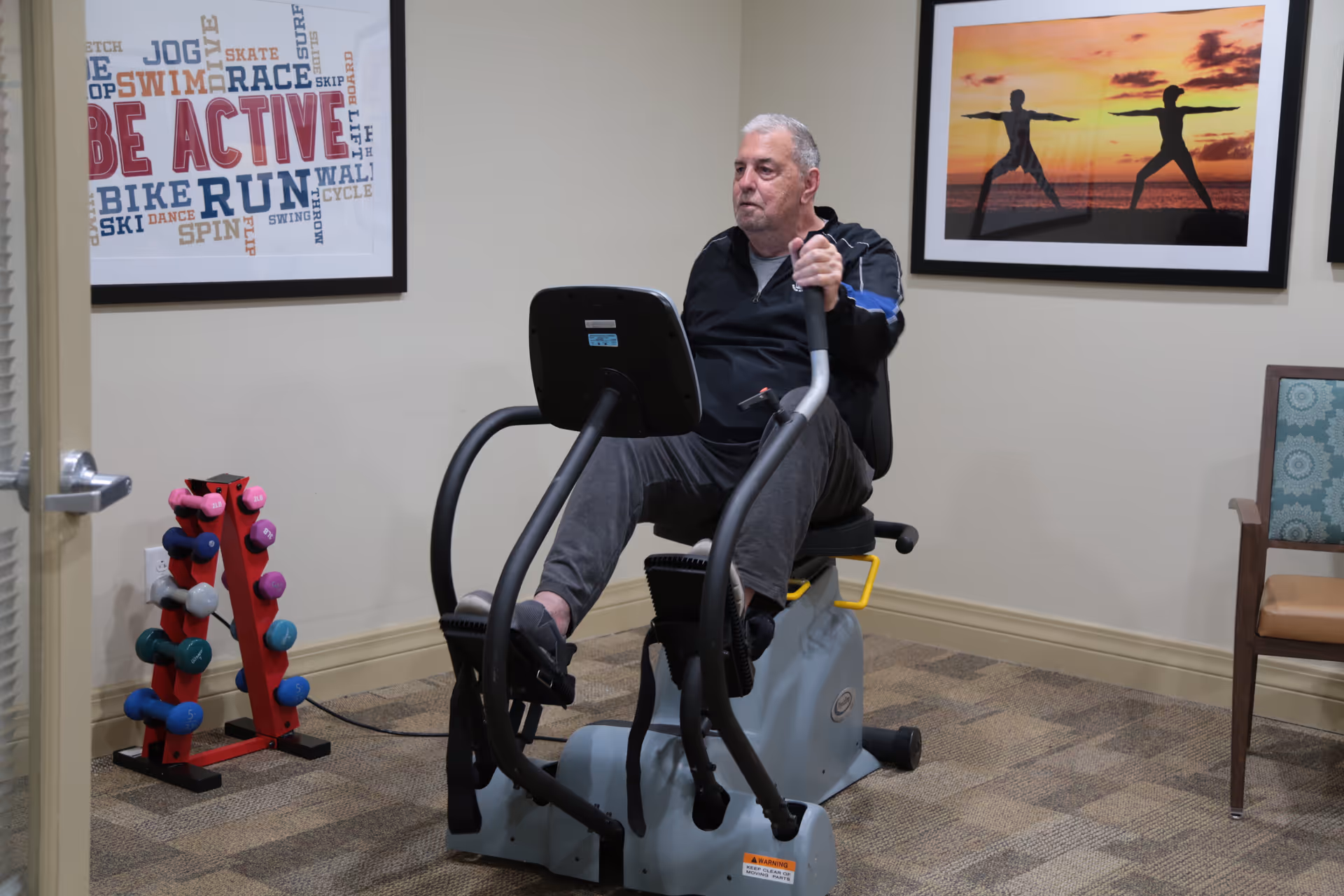 An elderly man exercising on a seated elliptical machine in a room with beige walls and carpeted floor. There is a rack of colorful dumbbells to the left and two framed pictures on the wall, one with the words 'BE ACTIVE' and various exercise-related terms, and the other showing two people doing yoga poses at sunset. A wooden chair with a patterned cushion is on the right side of the image.