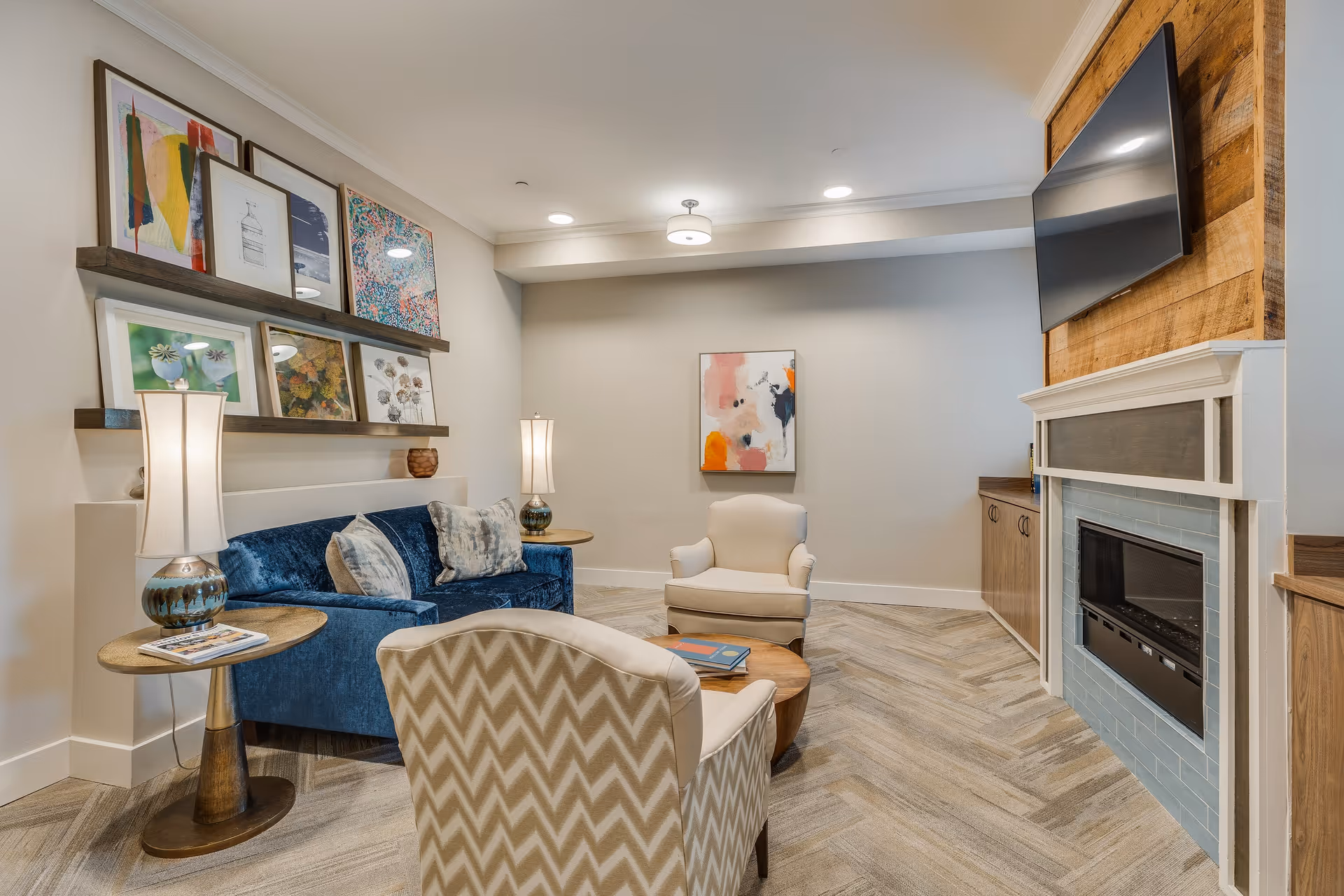 A cozy living room with a blue velvet sofa, two beige armchairs, a round wooden coffee table with books, two side tables with lamps, a fireplace with a mounted flat-screen TV above it, and colorful framed artwork on the walls.