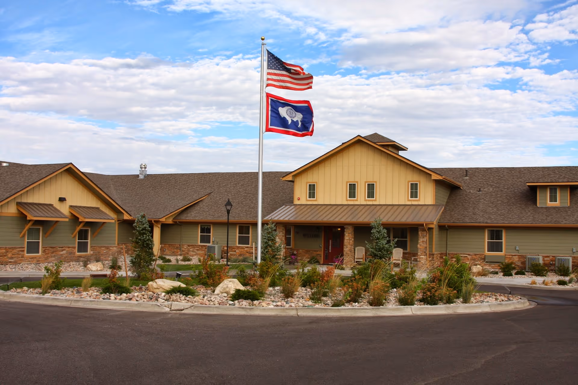 Front exterior of a single-story assisted living building with an American flag and a state flag flying above a landscaped roundabout.