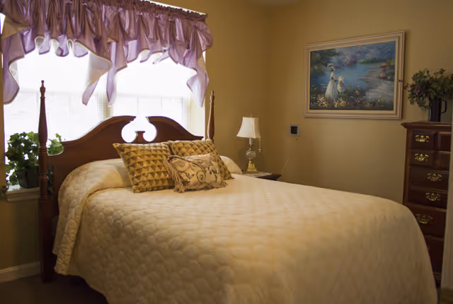 A neatly made bed with decorative pillows beneath a window with a lavender valance in a softly lit bedroom with a dresser and framed artwork.