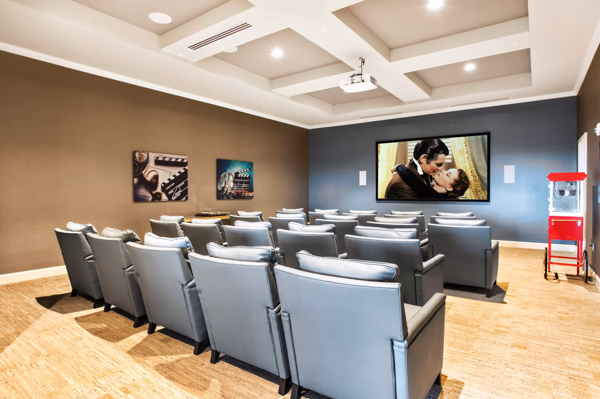 A small movie theater room with several rows of gray cushioned chairs facing a large screen showing a classic movie scene of a man and woman kissing. The room has beige walls with two film-themed pictures and a red popcorn machine in the corner.