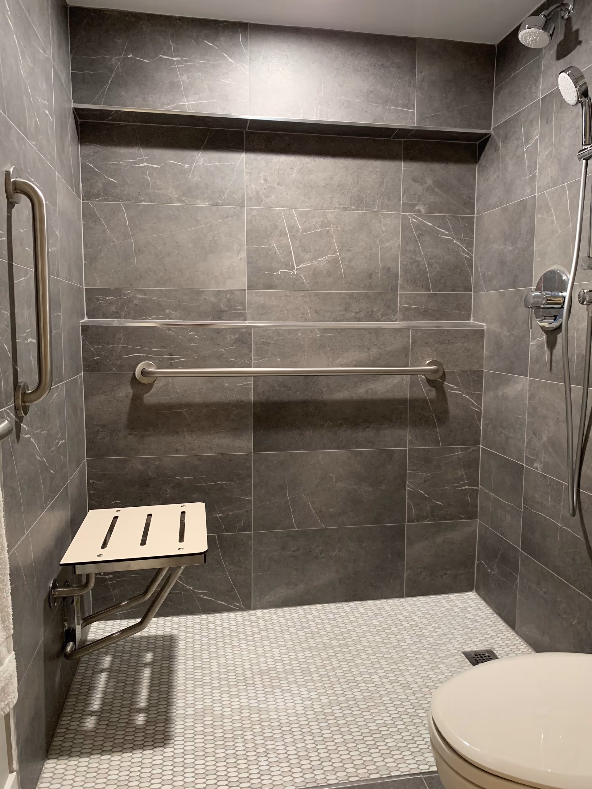 A modern accessible shower with gray tiled walls and a white tiled floor. The shower features a foldable seat, a horizontal grab bar, a vertical grab bar, and a handheld showerhead with controls mounted on the wall. A toilet is partially visible in the lower right corner.