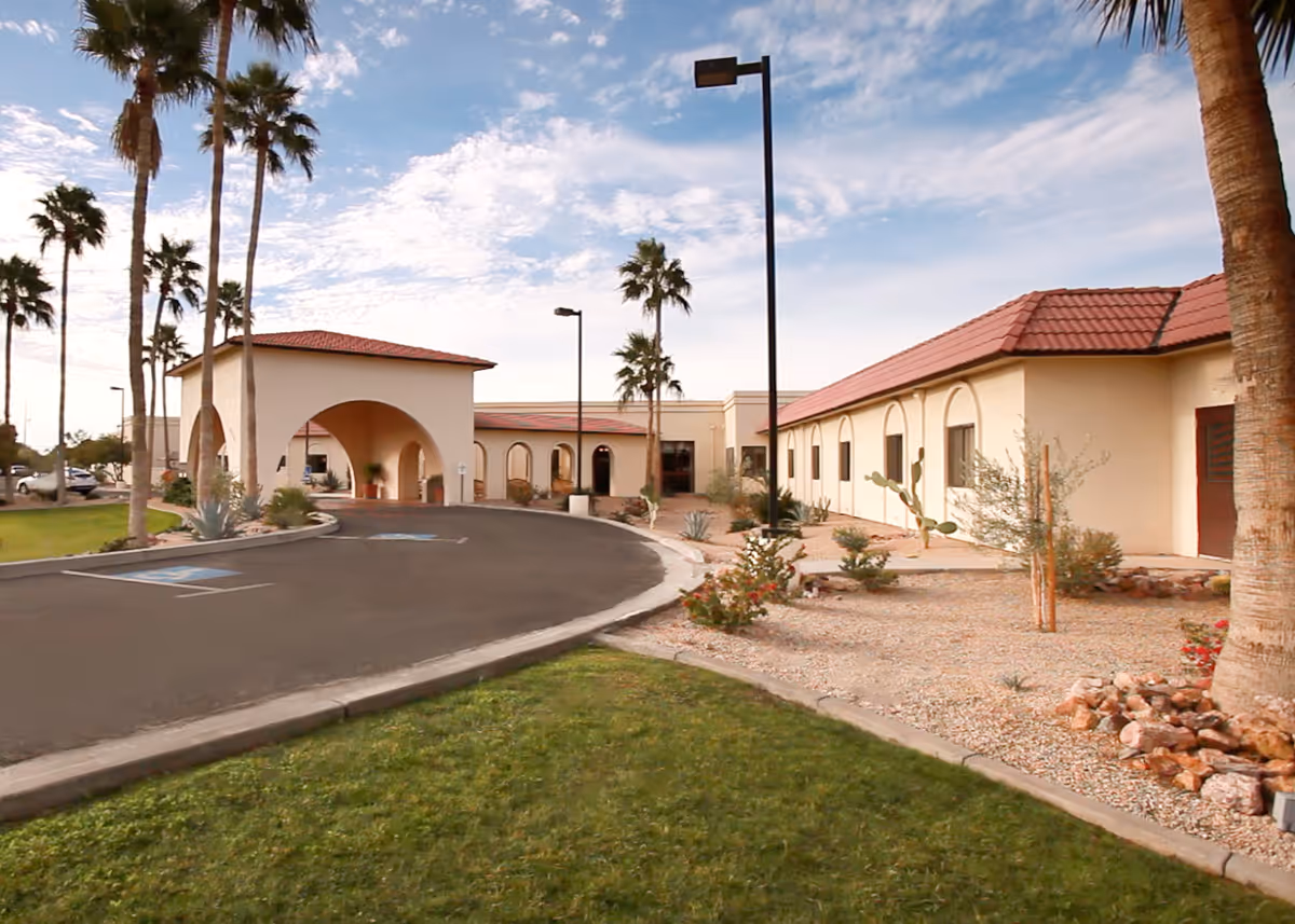 Exterior view of a single-story building with a red tile roof and arched entrance, surrounded by palm trees and desert landscaping under a partly cloudy sky.