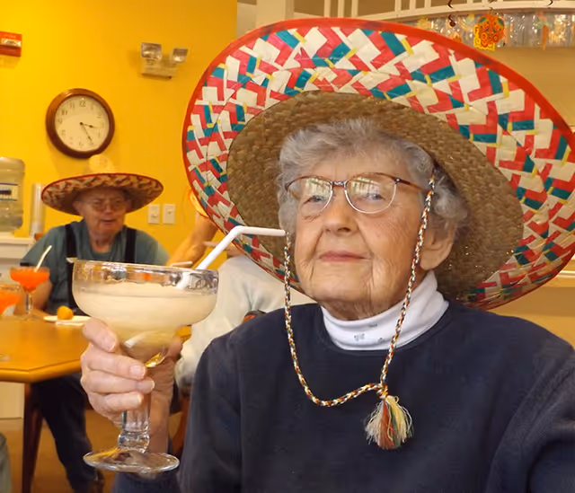 An elderly woman wearing glasses and a colorful sombrero hat is holding a large margarita glass with a straw, sitting at a table in a room with yellow walls. In the background, another elderly person also wearing a sombrero is seated at the table with drinks. A clock and water dispenser are visible on the wall.
