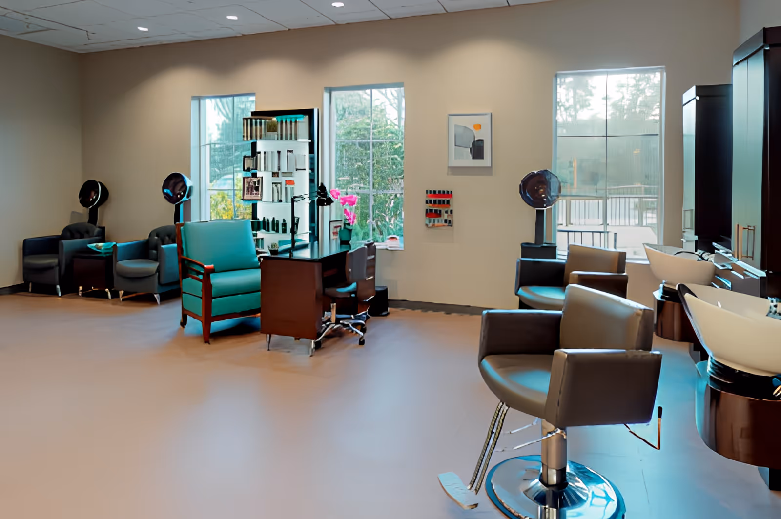 Interior view of a salon area in a senior living facility with multiple salon chairs, hair washing stations, hair dryers, and a manicure table. Large windows allow natural light to brighten the space, and shelves with hair care products are visible.