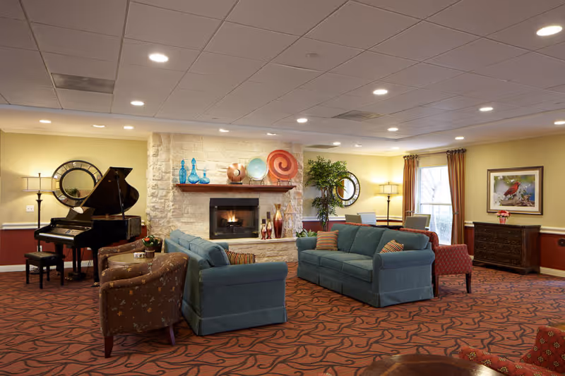 A cozy senior living facility common area with two blue sofas and two patterned armchairs arranged around a stone fireplace. Decorative vases and plates are displayed on the mantel. A grand piano is positioned in the corner next to a round mirror and a floor lamp. The room has warm yellow walls, red patterned carpet, and large windows with brown curtains letting in natural light.