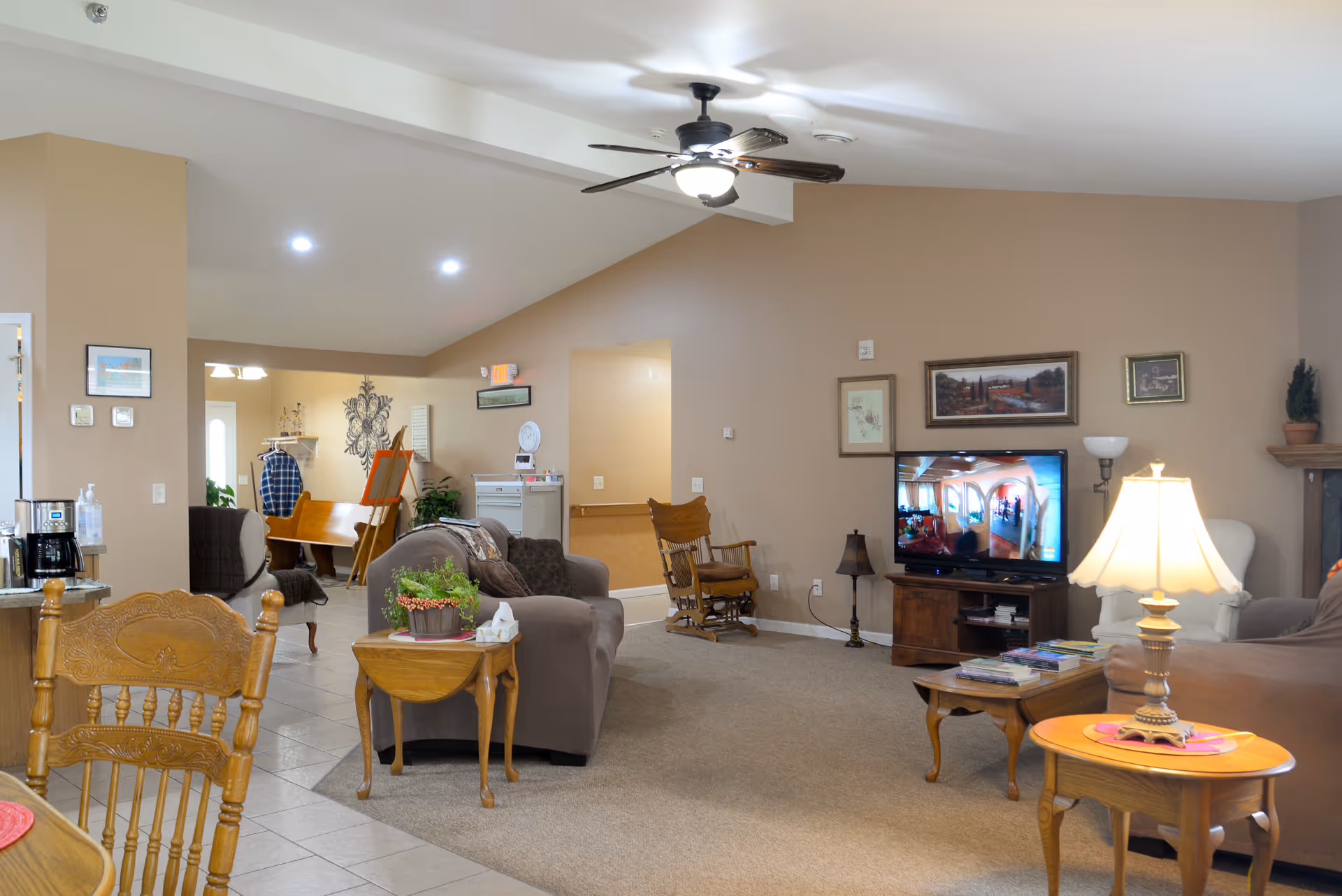 Spacious senior living community room with sofas, a TV, coffee tables, a rocking chair, and a dining area in the background.