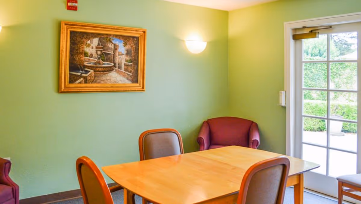 Small dining area with a wooden table and chairs, a framed painting on a green wall, and a glass door to the outside.
