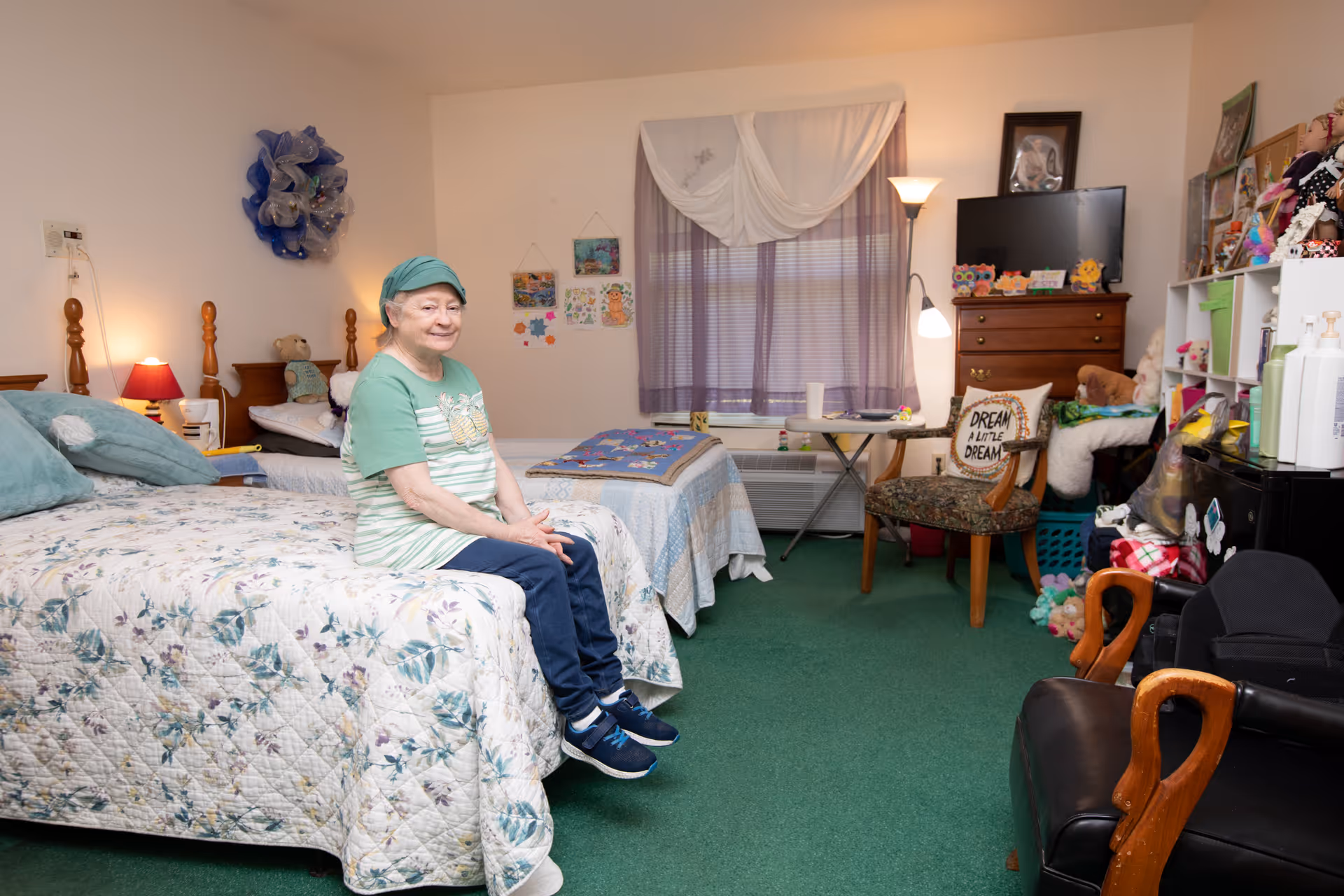 An elderly woman wearing a green headscarf and green striped shirt sits on the edge of a bed in a cozy room with green carpet. The room has two beds with floral and patchwork quilts, a wooden dresser with a TV on top, a chair with a pillow that says 'Dream a little dream', a small table, and shelves filled with various items and decorations. A window with purple curtains and a white sheer valance is in the background.