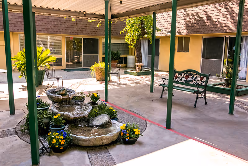 Covered courtyard with a tiered stone fountain, potted flowers, benches and patio chairs in front of sliding glass doors.