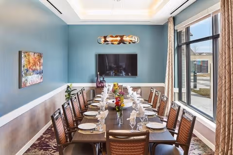 A dining room with a long rectangular table set for twelve people, featuring plates, glasses, napkins, and floral centerpieces. The room has blue walls with a white trim, a modern light fixture hanging above the table, a large window with beige curtains letting in natural light, and a flat-screen TV mounted on the wall.