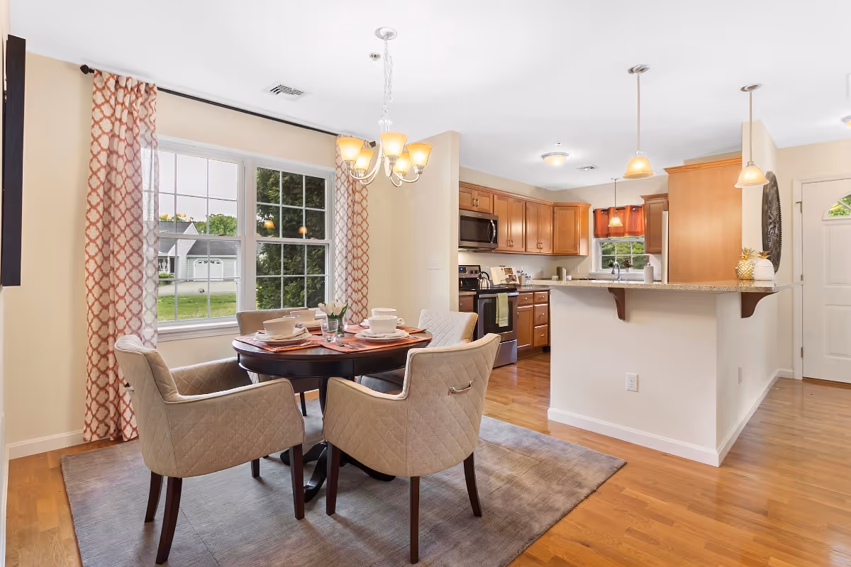 A bright dining area with a round wooden table set for four with beige upholstered chairs on a gray rug. The dining area is next to large windows with patterned curtains, and adjacent to a kitchen with wooden cabinets, stainless steel appliances, and a breakfast bar with pendant lights.