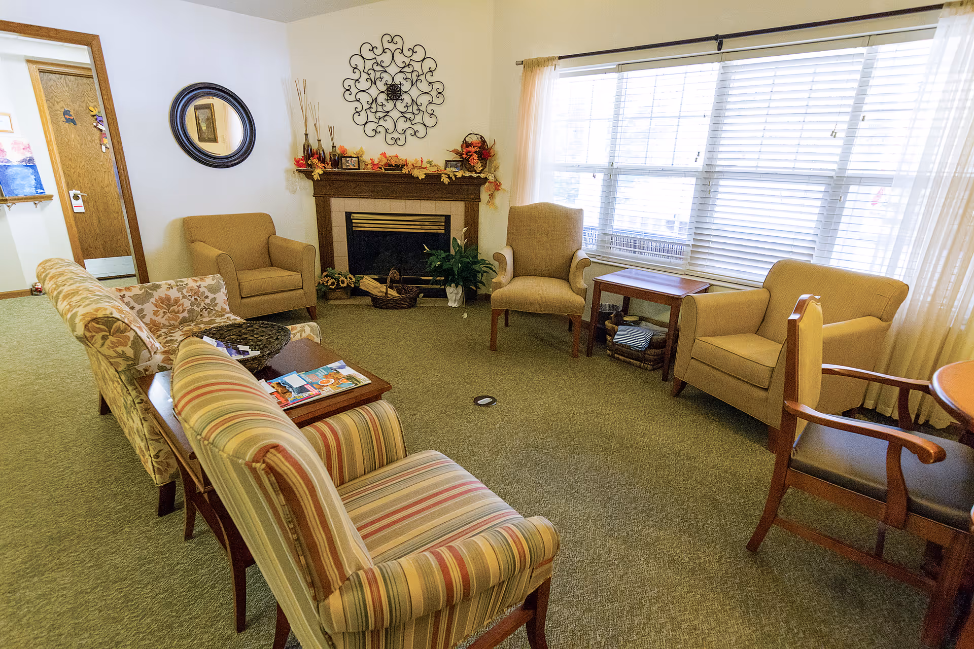 A cozy living room area with multiple upholstered chairs and a small sofa arranged around a wooden coffee table. There is a fireplace decorated with autumn leaves and vases, a round mirror on the wall, and a large window with blinds letting in natural light. The room has carpeted flooring and light-colored walls.