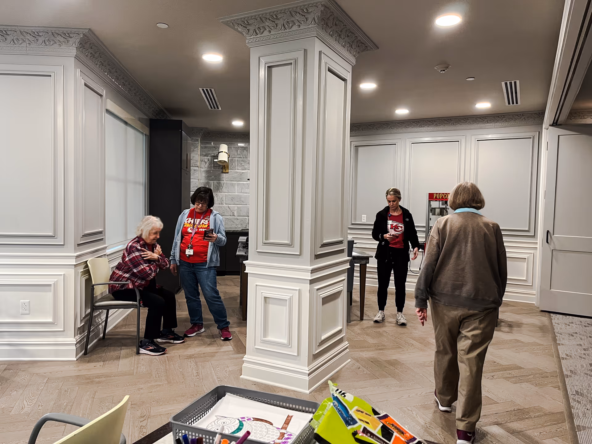 An indoor common area with four people, including two elderly women and two staff members wearing red Chiefs shirts. One elderly woman is seated while the others stand or walk around. The room features decorative white paneling, a popcorn machine, and a basket with games or activity supplies in the foreground.