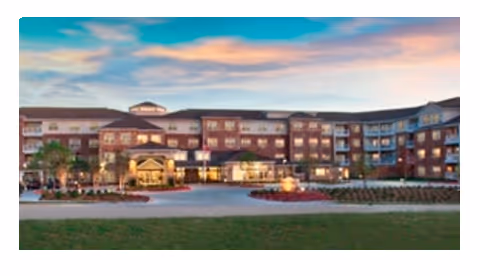 Front exterior of a multi-story brick senior living facility at dusk with a lit entrance and landscaped grounds.
