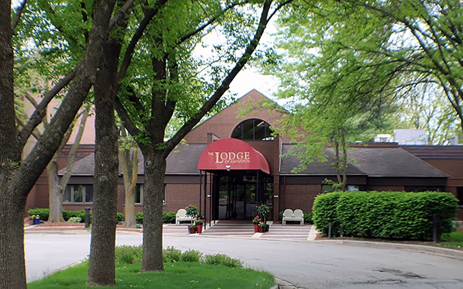 Front exterior view of a brick building with a red awning that reads 'The Lodge of Ashworth'. The entrance is framed by trees and greenery, with benches and potted plants near the doorway.