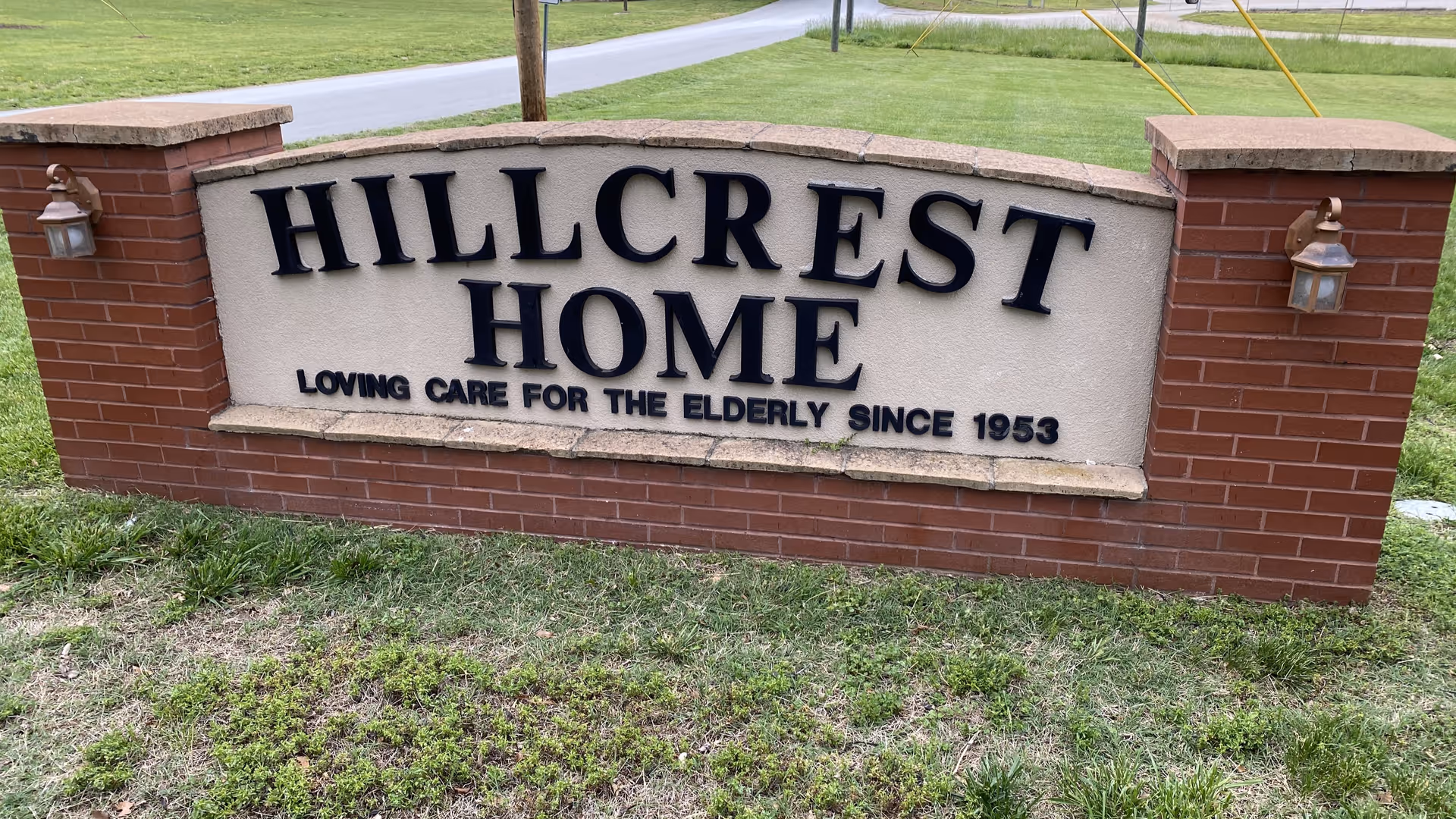 A brick and stone sign for Hillcrest Home with the text 'Loving Care for the Elderly Since 1953' displayed on a grassy area near a road.