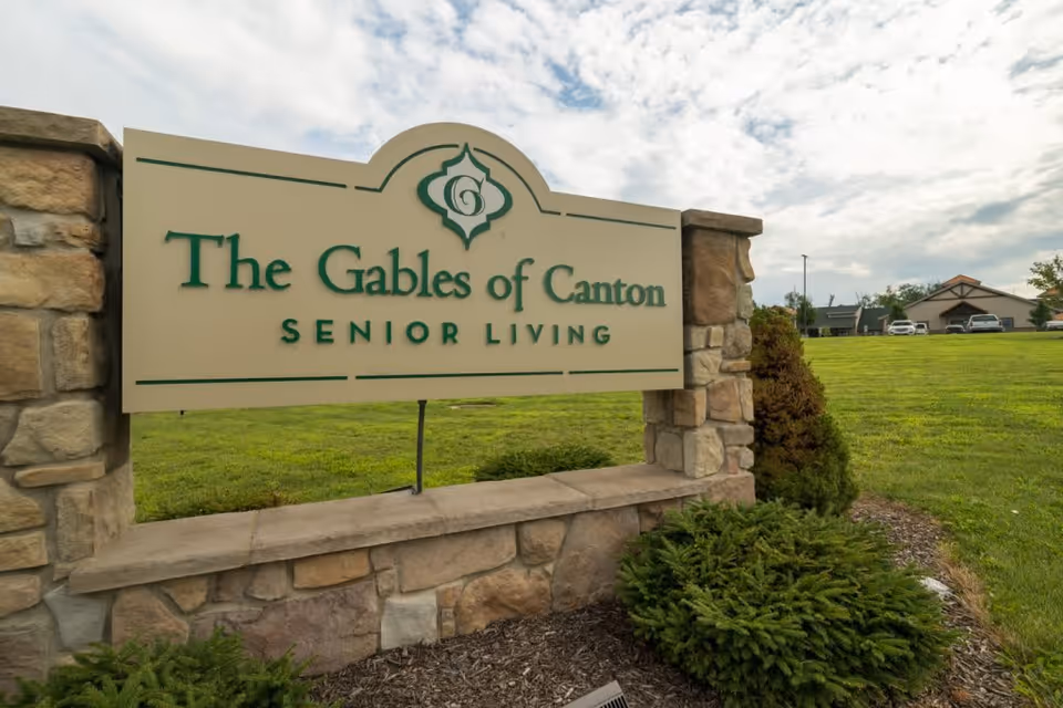 Stone and concrete sign for The Gables of Canton Senior Living set in a landscaped area with grass, bushes, and a partly cloudy sky in the background.