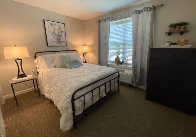 A cozy bedroom with a black metal bed frame, white bedding, and multiple pillows. There are two bedside tables each with a lamp, a window with gray curtains, a black dresser with decorative items on top, and a framed floral picture hanging on the wall.