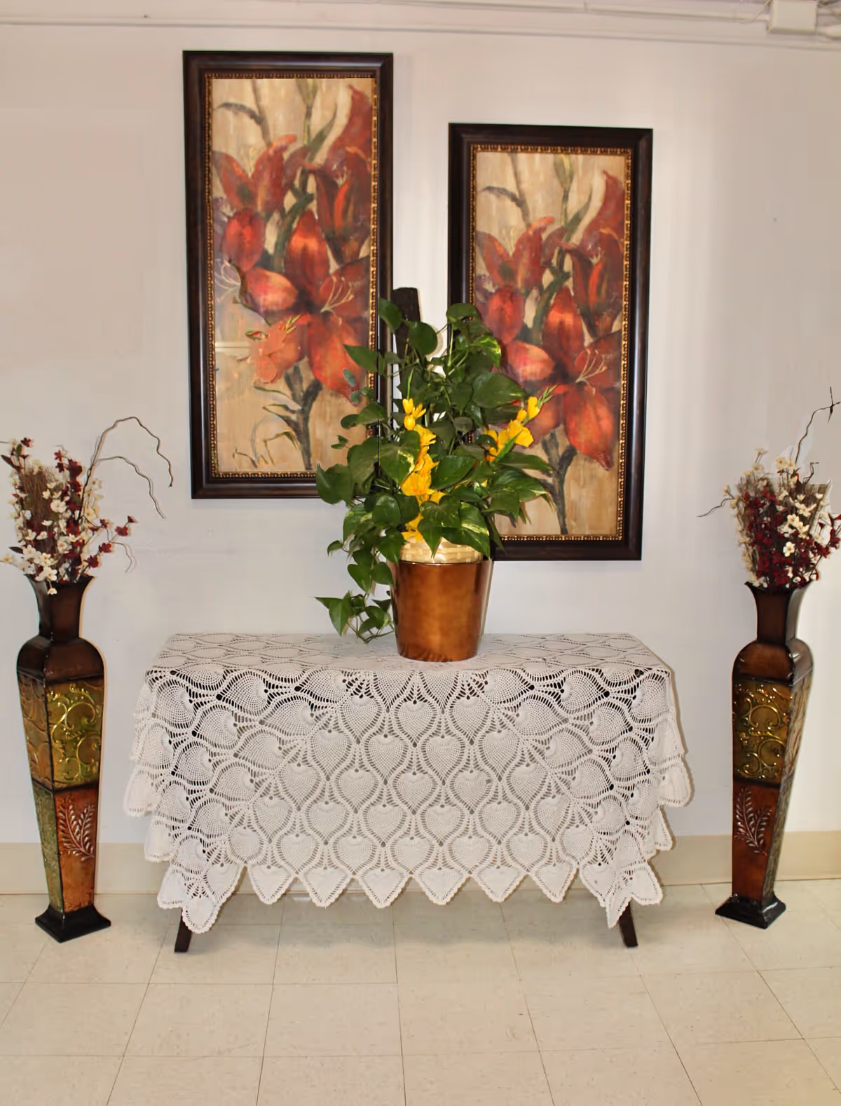 Interior entry table draped with a lace cloth topped by a potted plant, flanked by tall vases and two framed floral paintings.