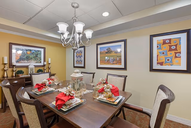 A dining room with a wooden table set for six people, featuring red napkins, decorative plates, glasses, and a centerpiece lantern. The room has beige walls adorned with four framed pictures and a chandelier hanging above the table. There is a sideboard with candles and decorative items against one wall.