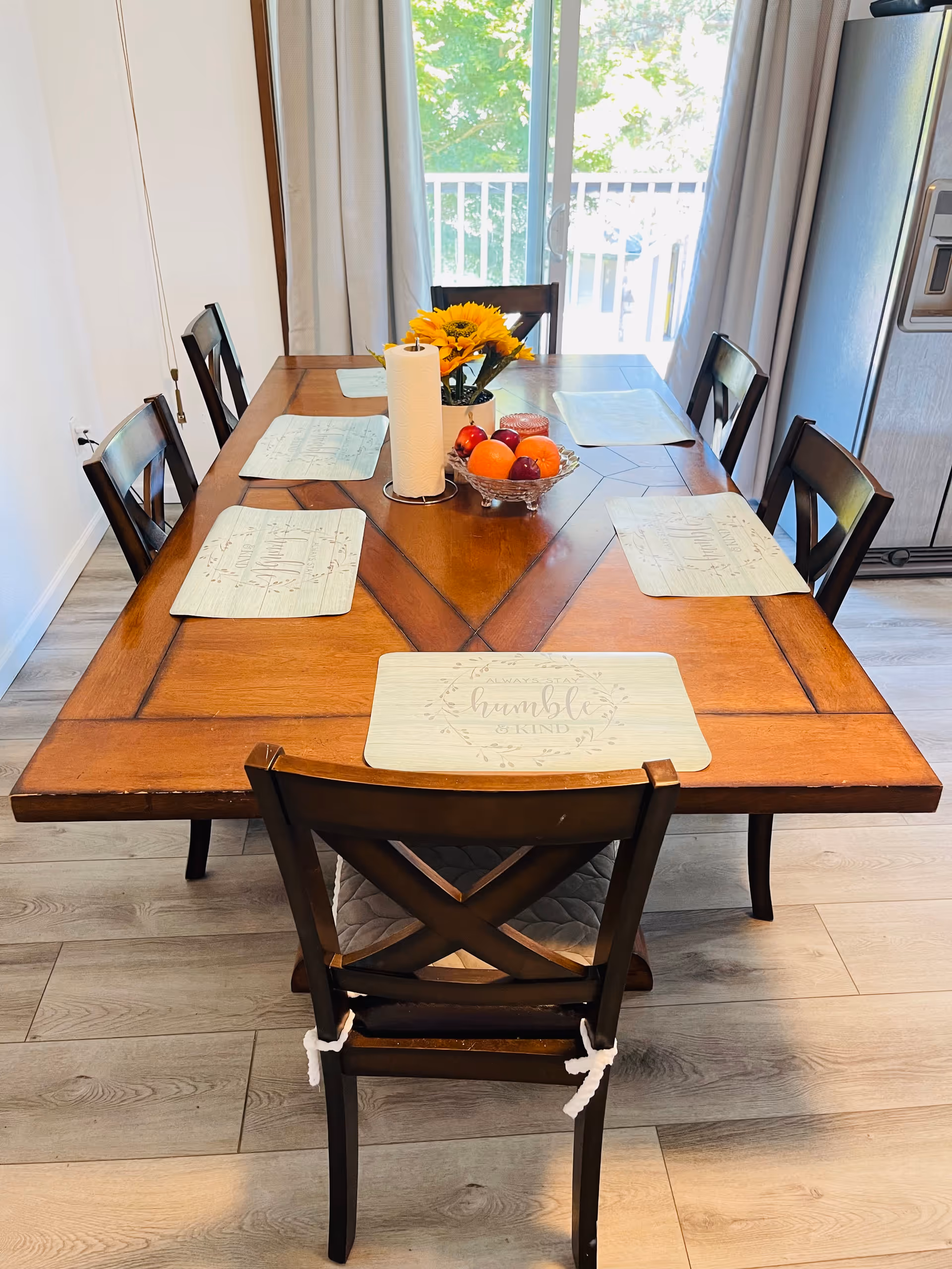 A wooden dining table set with six chairs in a bright room. The table has six placemats, a paper towel roll, a bowl of fruit, and a vase with sunflowers. Behind the table is a sliding glass door with curtains, letting in natural light. A stainless steel refrigerator is visible to the right.