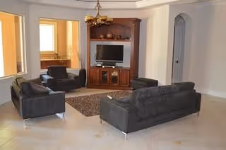 Spacious living room with three dark gray sofas and an armchair arranged around a rug facing a wooden entertainment center with a TV, tile floors, chandelier, and arched doorways.