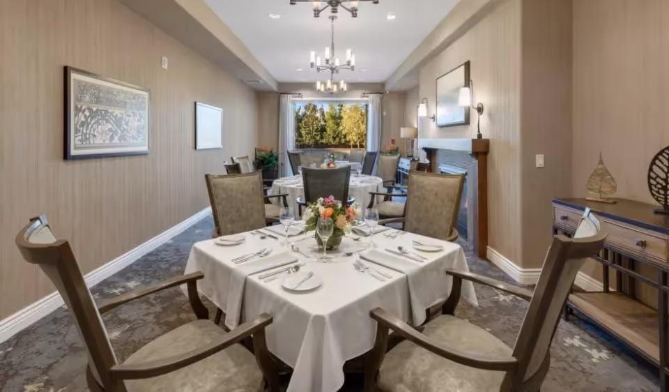 Well-appointed dining room with white-clothed tables set for a meal, upholstered chairs, chandeliers, and a window showing trees.