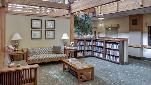 A cozy senior living common area featuring wooden framed sofas with patterned cushions, a wooden coffee table with books, two table lamps on side tables, framed artwork on the wall, a bookshelf filled with books, and a large potted plant. The space has a warm, inviting atmosphere with wooden beams and soft lighting.