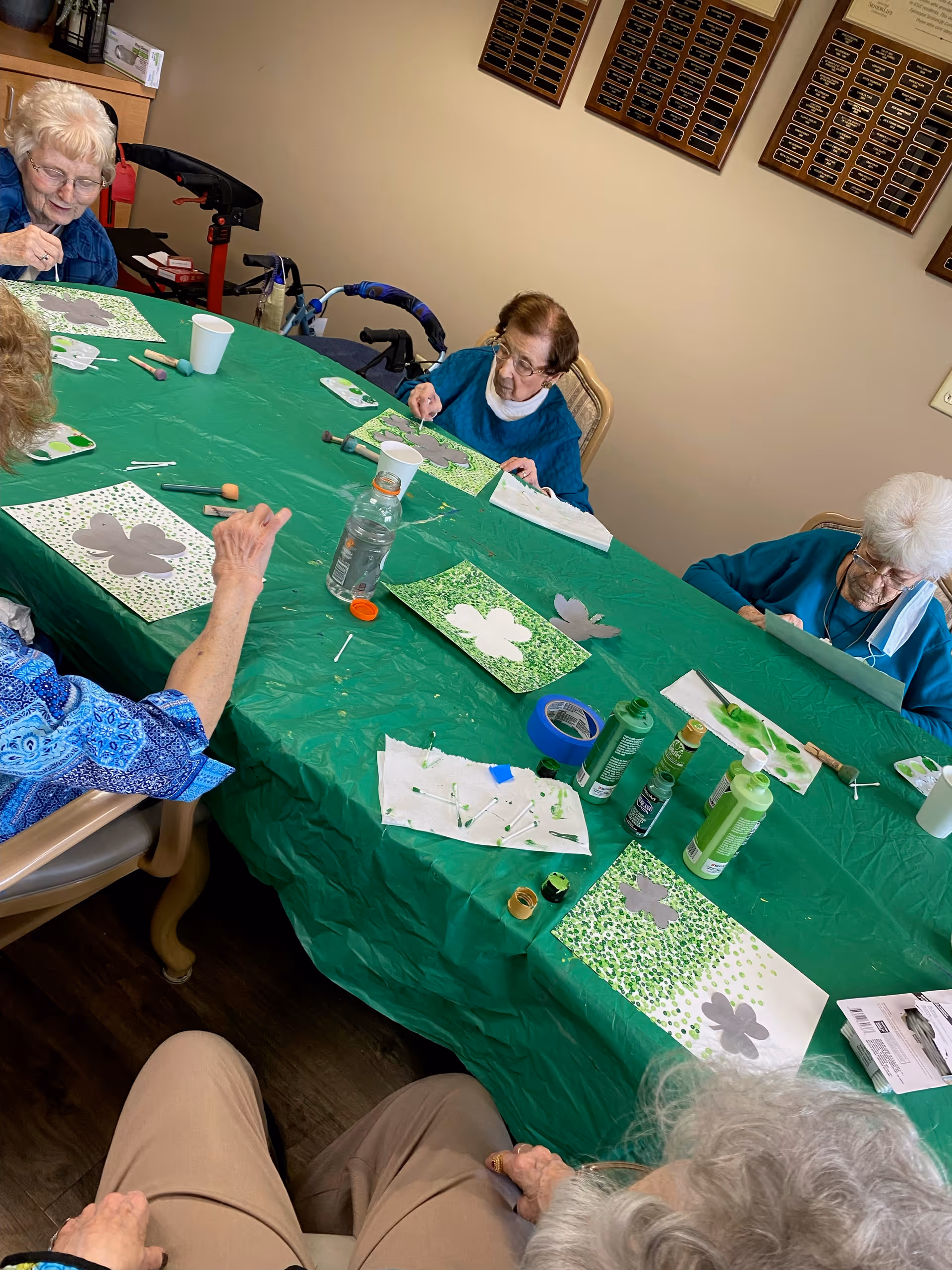 A group of elderly women seated around a table covered with a green plastic tablecloth, engaged in a painting activity using stencils shaped like shamrocks. Various art supplies including paint bottles, brushes, tape, and paper cups are scattered on the table. The room has beige walls with plaques hanging on them.
