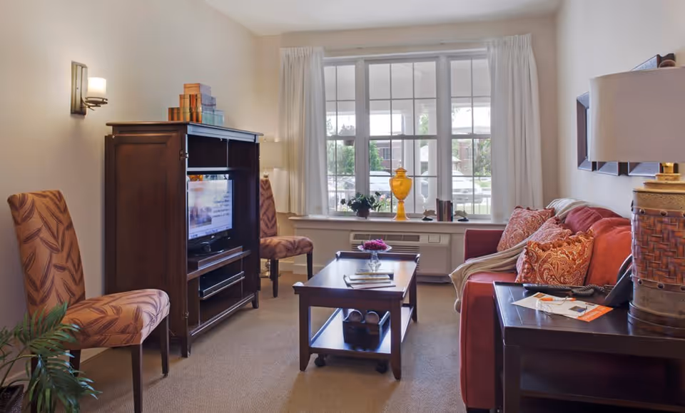 A cozy living room with a red sofa adorned with patterned cushions and a throw blanket. There is a wooden coffee table with a small flower vase and books on it. Two patterned chairs are placed near a wooden TV cabinet with a television inside. A large window with white curtains allows natural light to brighten the room. A table lamp and some decorative items are also visible.