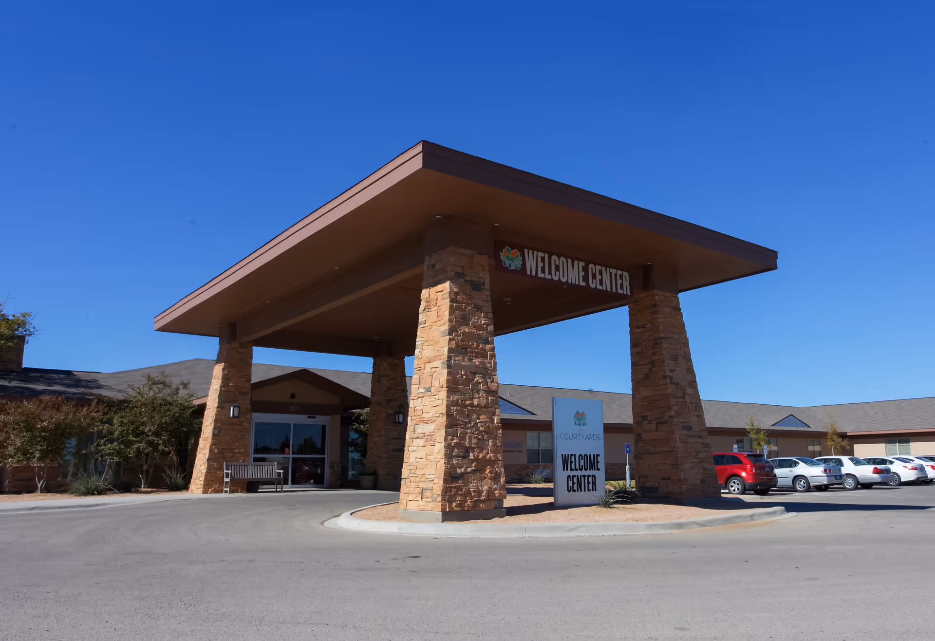 Drive-through entrance with stone pillars and 'Welcome Center' signage at an assisted living building under a clear blue sky.