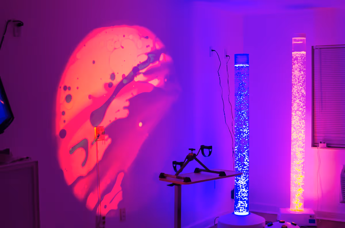A sensory room with colorful lighting effects including a large red and purple light projection on the wall, two tall bubble tubes emitting blue and pink light, and a small exercise pedal machine on a stand.
