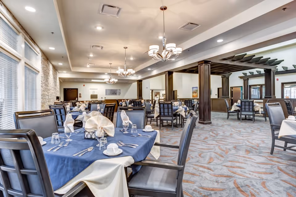 Spacious dining room with multiple neatly set tables, blue tablecloths, upholstered chairs, chandeliers and large windows.