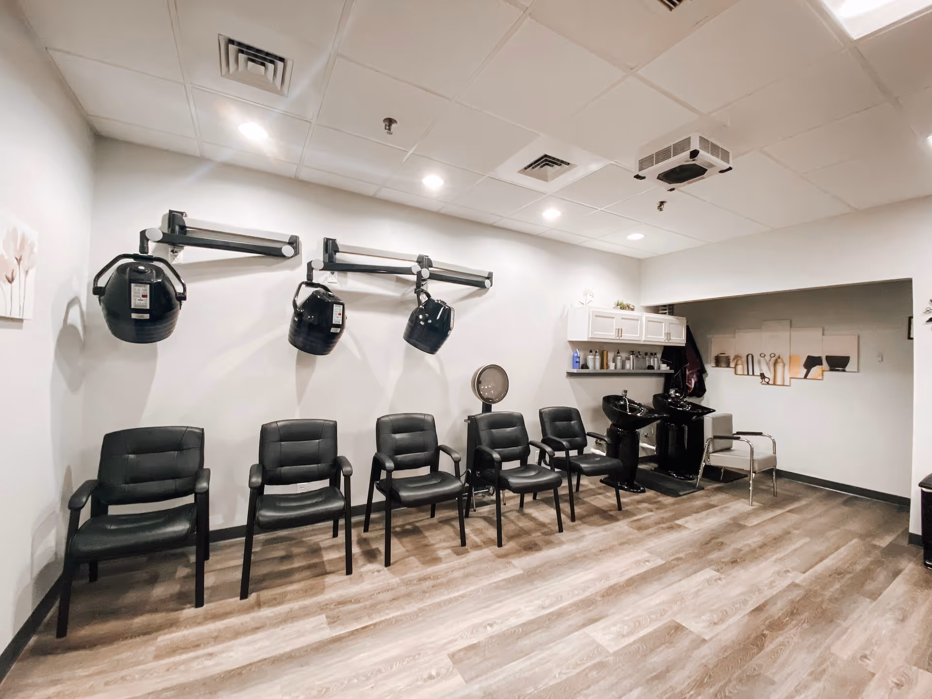 Interior of a salon area with five black chairs lined up against a wall. Above three of the chairs are black hair drying machines mounted on the wall. To the right, there are two black hair washing sinks with shelves above holding various hair care products. The floor is wood-patterned, and the ceiling has recessed lighting and ventilation.
