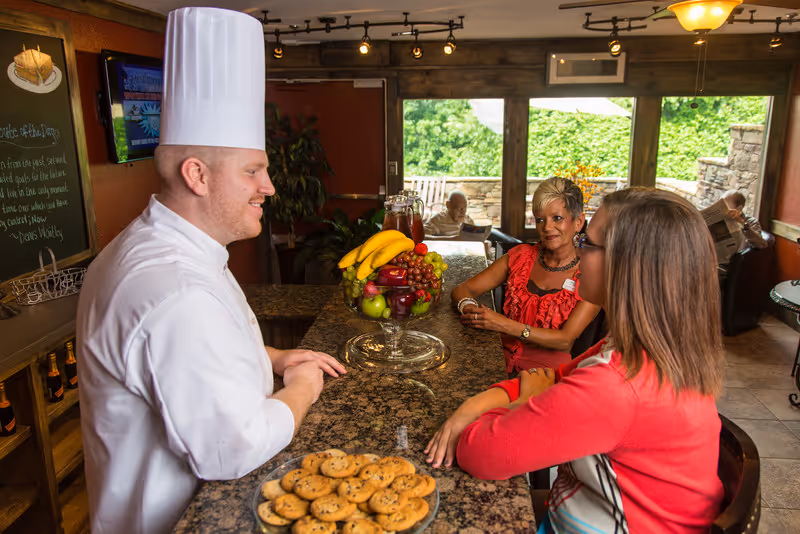 A chef in a white uniform and tall hat stands behind a granite counter with a plate of cookies and a large bowl of fruit. Two women sit on the other side of the counter, engaged in conversation with the chef. In the background, two elderly people are seated near large windows with a view of greenery outside.