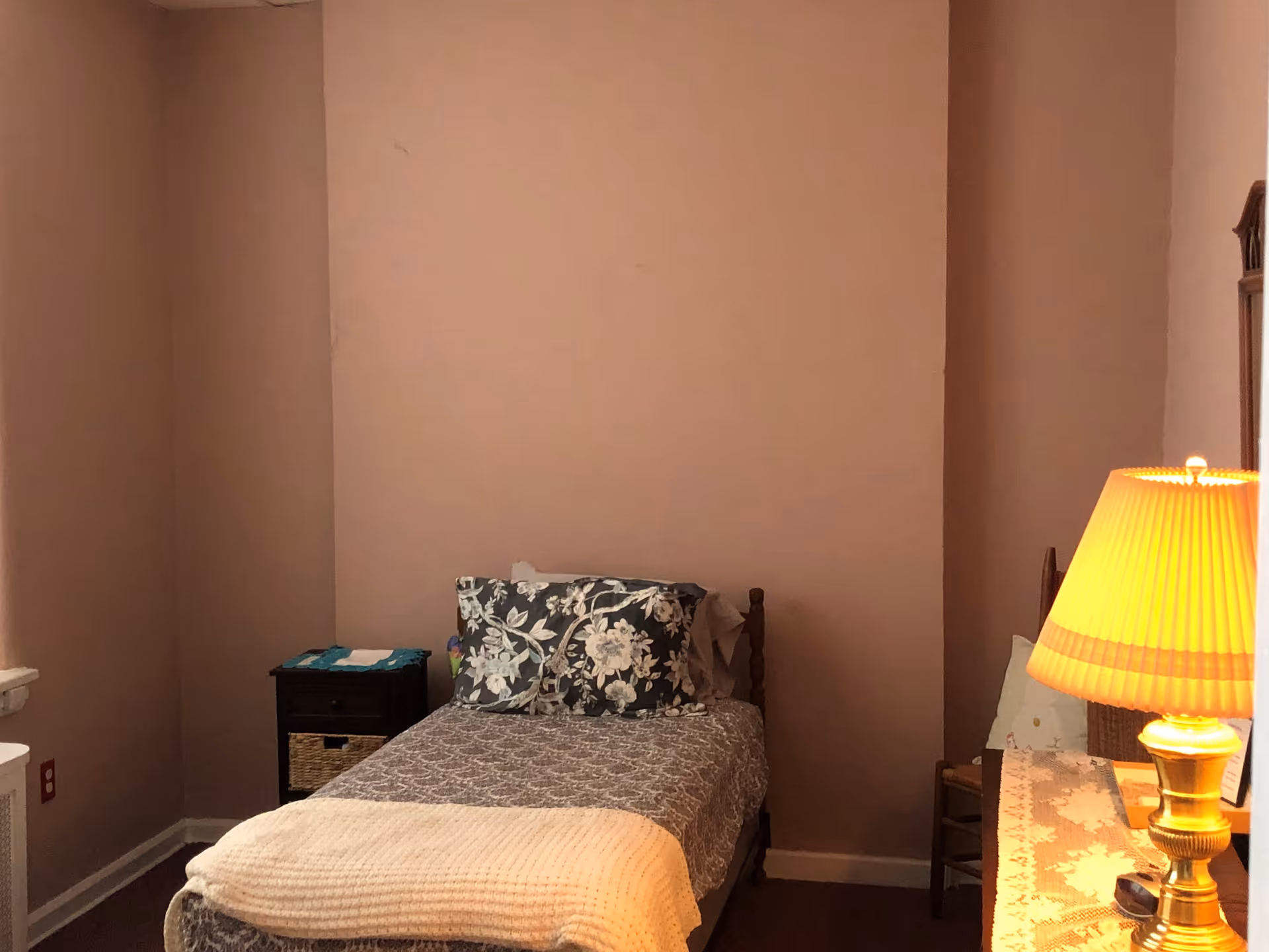 A small bedroom with a single bed against a plain beige wall. The bed has a patterned bedspread, floral pillows, and a cream-colored blanket at the foot. To the left of the bed is a small black nightstand with a basket and some items on top. On the right side, there is a wooden chair and a dresser with a lace runner and a lit table lamp with a pleated shade.