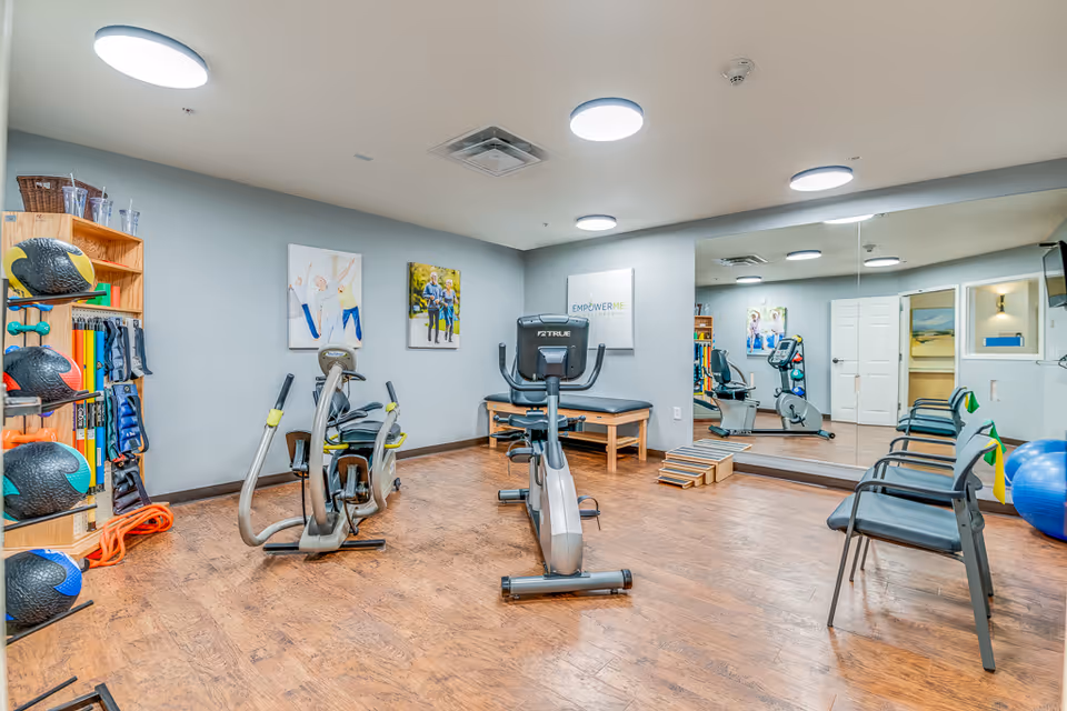 A bright and clean exercise room with wooden flooring, featuring stationary exercise bikes, a rack with medicine balls and resistance bands, chairs lined up against the wall, a large mirror, and motivational posters on the walls.