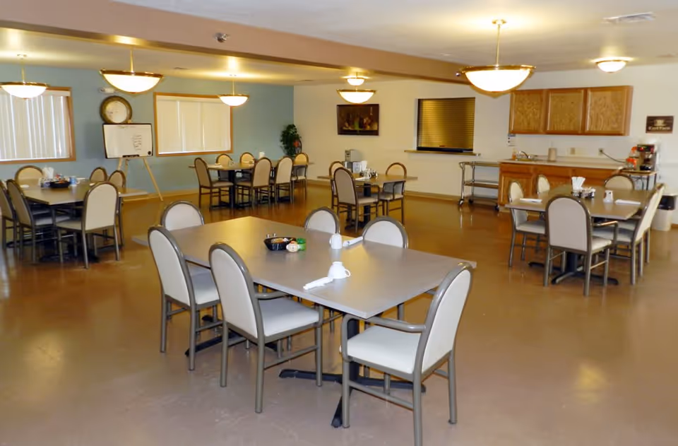 A dining room in an assisted living facility with multiple tables and chairs arranged neatly. The room has a light blue and white color scheme with overhead pendant lights. There is a clock and a whiteboard on one wall, a serving counter with cabinets, and a coffee station on the right side.