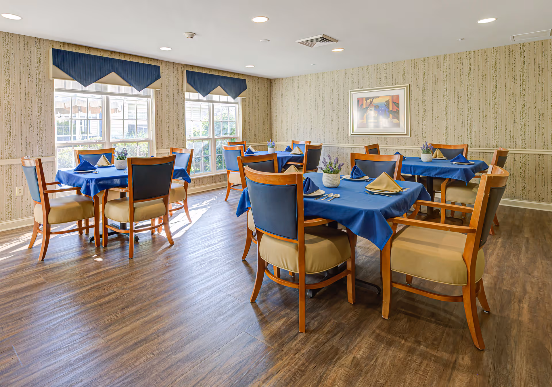 A dining room with several wooden tables covered with blue tablecloths and set with folded napkins and silverware. Each table is surrounded by wooden chairs with beige cushions and blue backs. The room has large windows with blue valances, patterned wallpaper, a framed picture on the wall, and wood flooring.